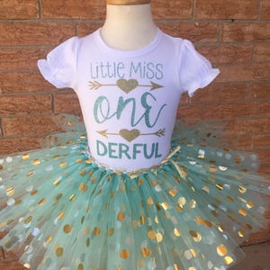 May include: A white t-shirt with the text "Little Miss one DERFUL" in gold and teal glitter. The shirt is paired with a teal tulle skirt with gold polka dots.