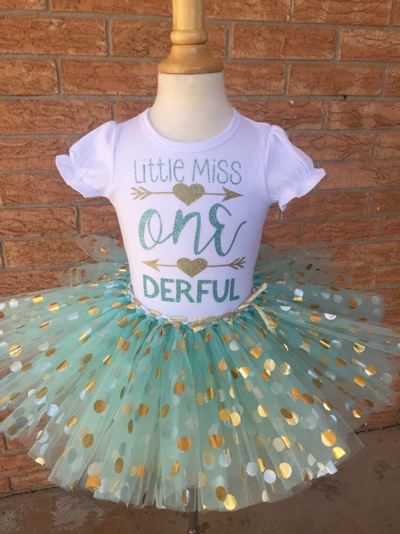 May include: A white t-shirt with the text "Little Miss one DERFUL" in gold and teal glitter. The shirt is paired with a teal tulle skirt with gold polka dots.