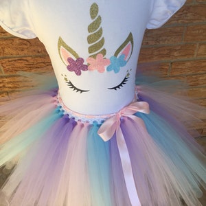 Peut inclure: Un t-shirt blanc avec un motif de licorne comprenant une corne dor&eacute;e paillet&eacute;e, des oreilles roses et une couronne de fleurs roses, violettes et bleues. Le t-shirt est associ&eacute; &agrave; un tutu en tulle arc-en-ciel avec un ruban de satin rose.