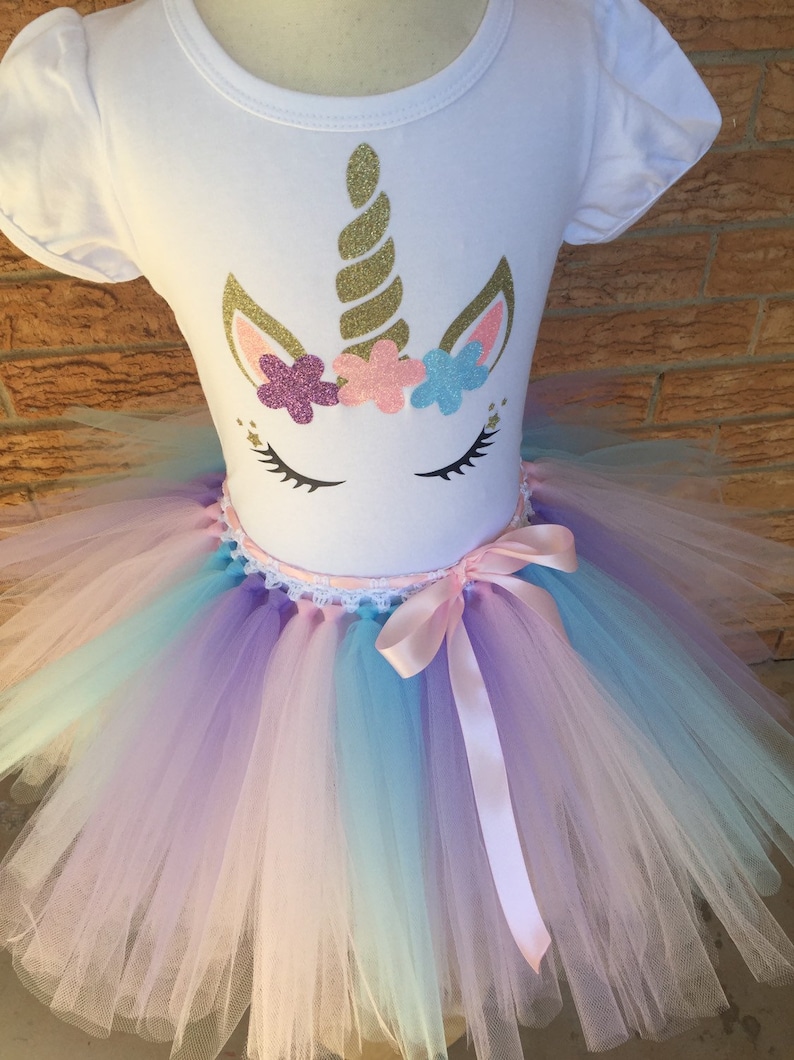 Peut inclure: Un t-shirt blanc avec un motif de licorne comprenant une corne dor&eacute;e paillet&eacute;e, des oreilles roses et une couronne de fleurs roses, violettes et bleues. Le t-shirt est associ&eacute; &agrave; un tutu en tulle arc-en-ciel avec un ruban de satin rose.