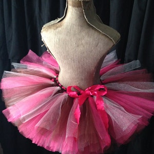 May include: A pink and brown tulle tutu with a pink satin bow. The tutu is on a dress form.