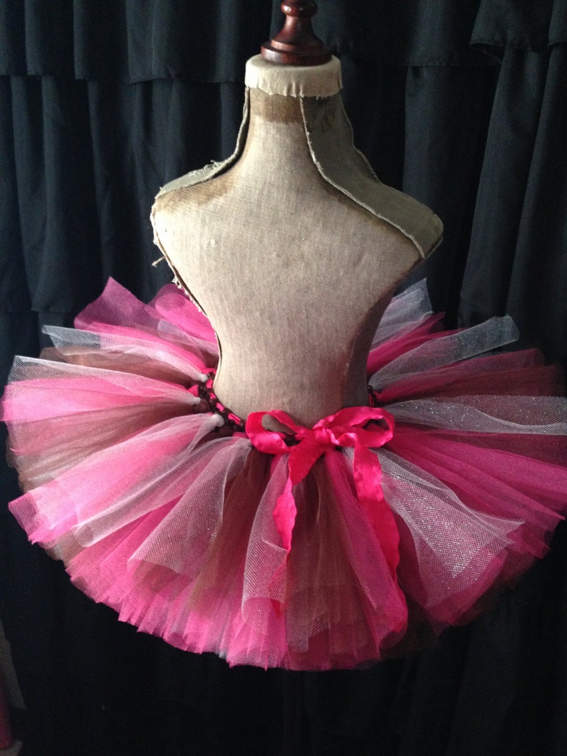 May include: A pink and brown tulle tutu with a pink satin bow. The tutu is on a dress form.