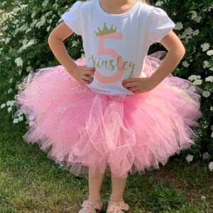 May include: A young girl wearing a white t-shirt with a gold crown and the number 5, the name Kinsley, and pink glitter. She is also wearing a pink tulle tutu with gold glitter.
