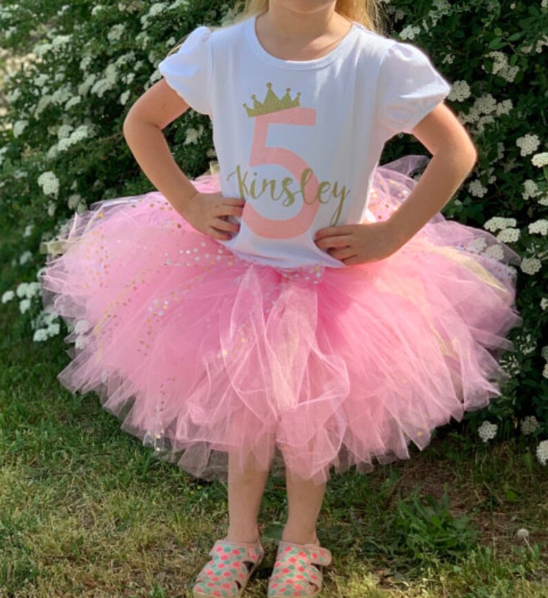 May include: A young girl wearing a white t-shirt with a gold crown and the number 5, the name Kinsley, and pink glitter. She is also wearing a pink tulle tutu with gold glitter.