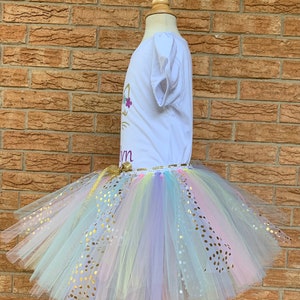 May include: A white t-shirt with a gold unicorn design and the name "Lynn" on it. The shirt is paired with a rainbow tulle tutu with gold glitter dots.
