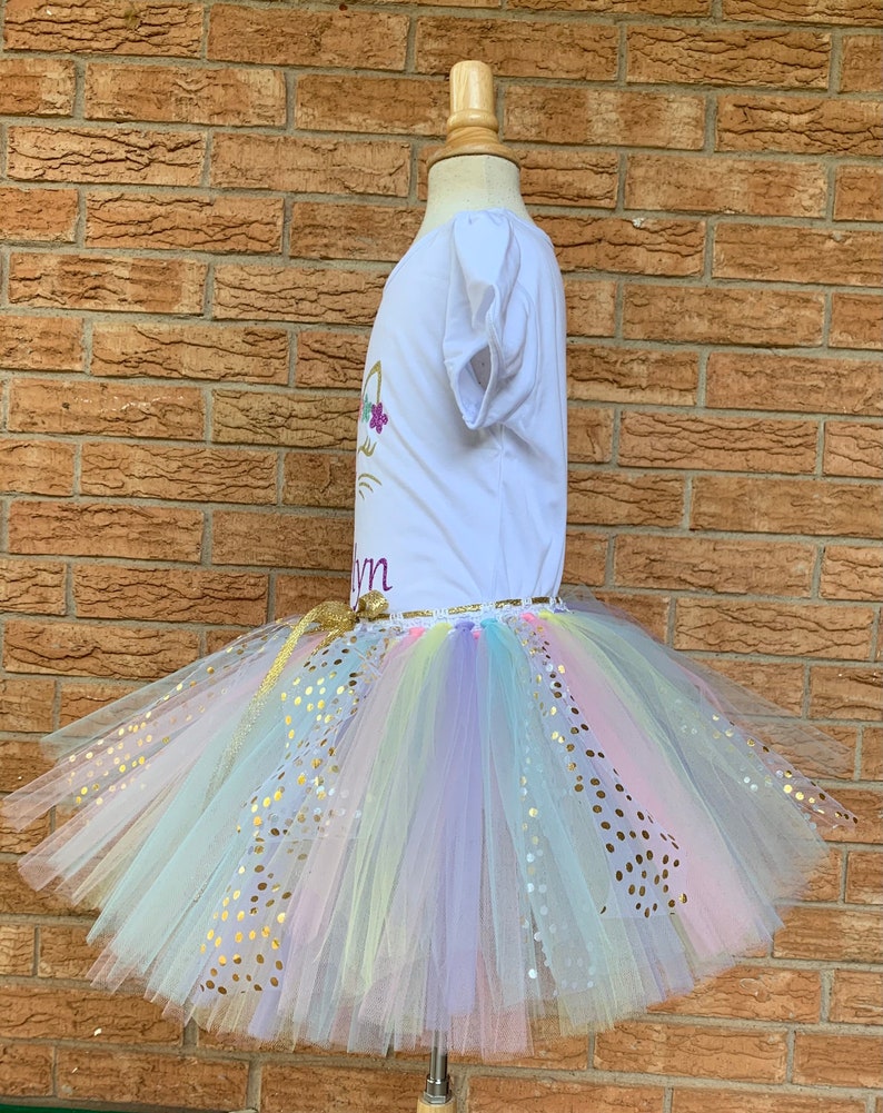 May include: A white t-shirt with a gold unicorn design and the name "Lynn" on it. The shirt is paired with a rainbow tulle tutu with gold glitter dots.