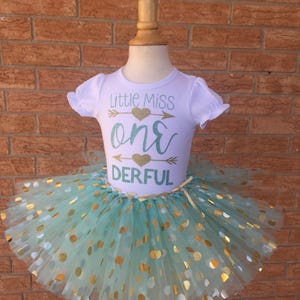 May include: A white t-shirt with the text "Little Miss One-derful" in gold glitter with a green heart and arrow. The shirt is paired with a mint green tulle tutu with gold polka dots.