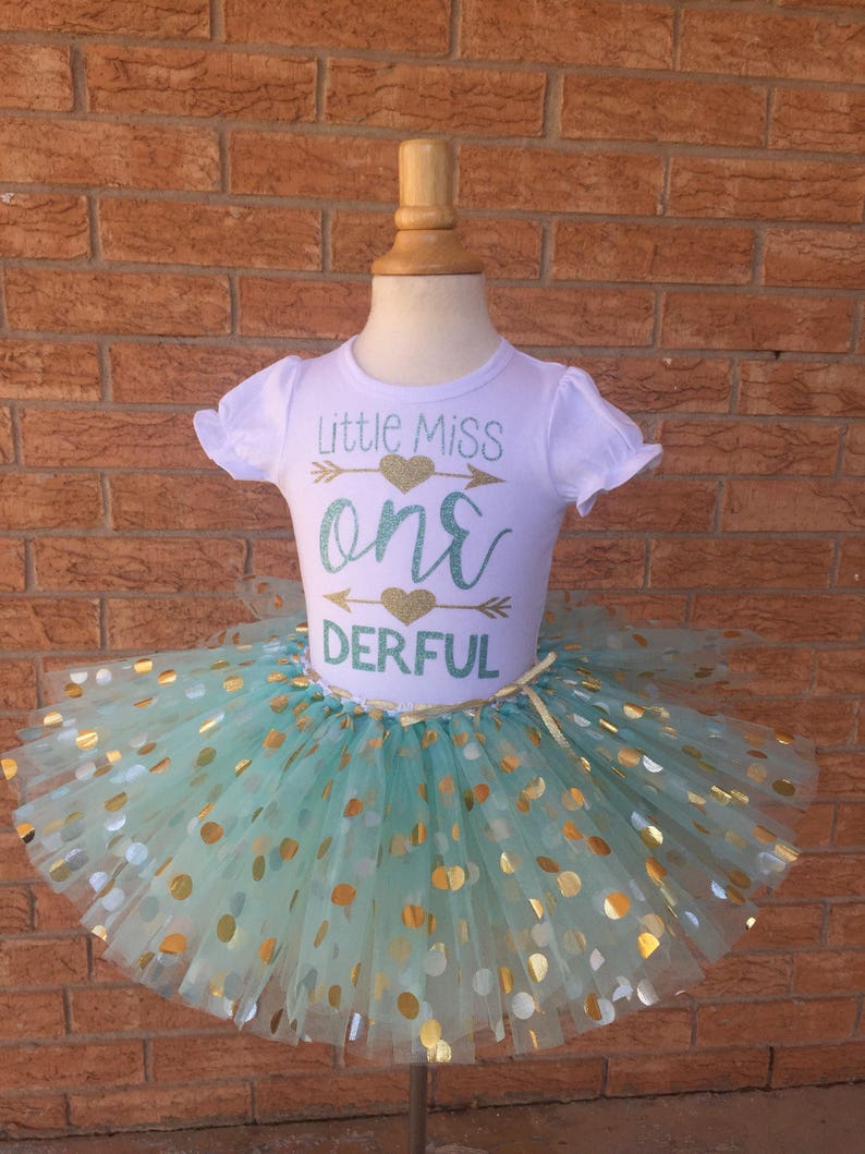 May include: A white t-shirt with the text "Little Miss One-derful" in gold glitter with a green heart and arrow. The shirt is paired with a mint green tulle tutu with gold polka dots.