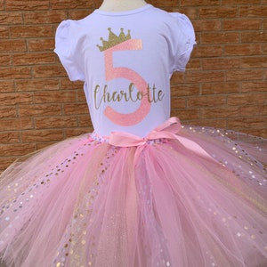 May include: A pink tulle tutu with gold glitter polka dots and a pink satin ribbon. A white t-shirt with a gold crown and the number 5 in pink glitter with the name Charlotte in gold glitter.