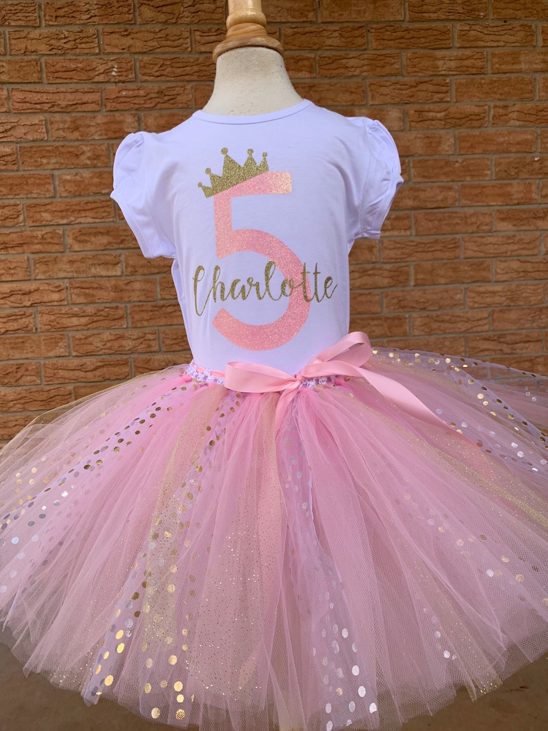 May include: A pink tulle tutu with gold glitter polka dots and a pink satin ribbon. A white t-shirt with a gold crown and the number 5 in pink glitter with the name Charlotte in gold glitter.