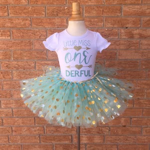 May include: A white t-shirt with the text "Little Miss One Derful" in gold glitter lettering and a gold glitter heart with arrows on either side. The shirt is paired with a mint green tulle tutu with gold polka dots.