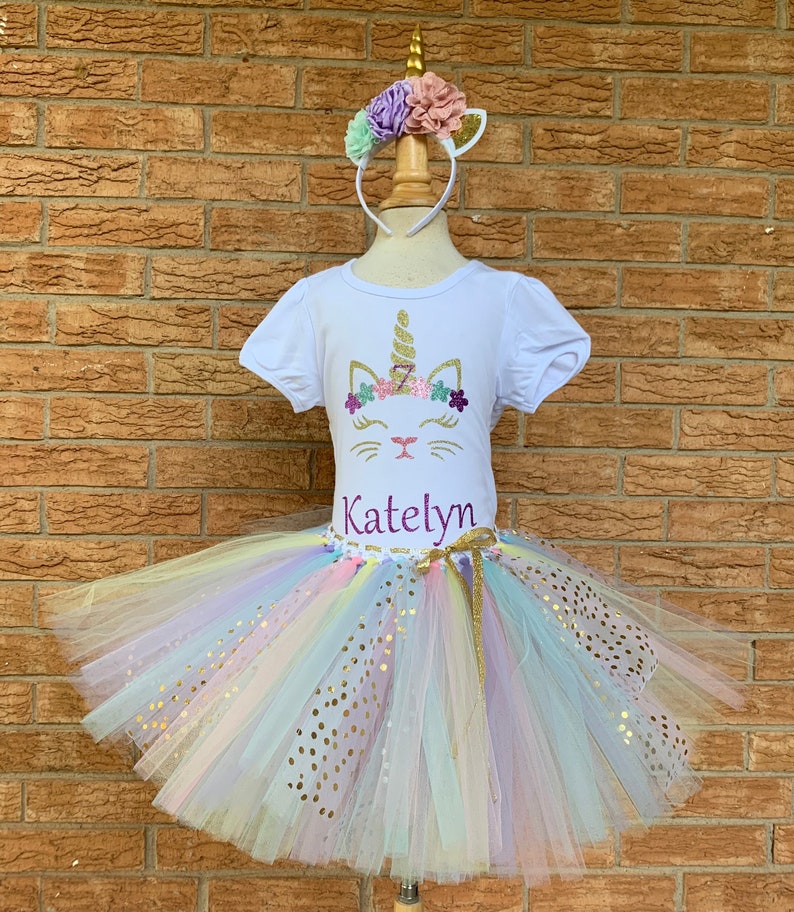 May include: A white t-shirt with a unicorn cat design and the name "Katelyn" in purple glitter. The shirt is paired with a rainbow tulle tutu with gold glitter polka dots. A unicorn headband with pink, purple, and green flowers is included.