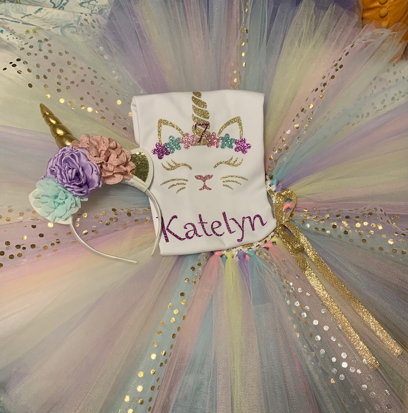 May include: A white tulle tutu with gold glitter dots and a rainbow ombre effect. A white t-shirt with a unicorn cat design and the name "Katelyn" is on top of the tutu. A unicorn headband with purple, pink, and blue flowers is on the left side of the tutu.