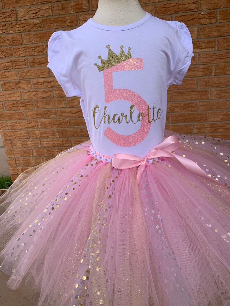 May include: A pink tulle tutu skirt with gold glitter polka dots. A white t-shirt with a pink number 5 and a gold glitter crown on top of the number. The name Charlotte is written in cursive below the number.