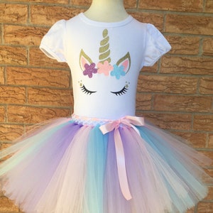 Peut inclure: Un t-shirt blanc avec un motif de licorne comprenant une corne dor&eacute;e paillet&eacute;e, des fleurs roses, violettes et bleues, et des yeux ferm&eacute;s. Le t-shirt est associ&eacute; &agrave; une jupe tutu en tulle multicolore avec un ruban de satin rose.