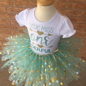 May include: A white t-shirt with the text "Little Miss One Derful" in gold glitter and a green arrow with a gold heart. The shirt is paired with a mint green tulle tutu with gold polka dots.