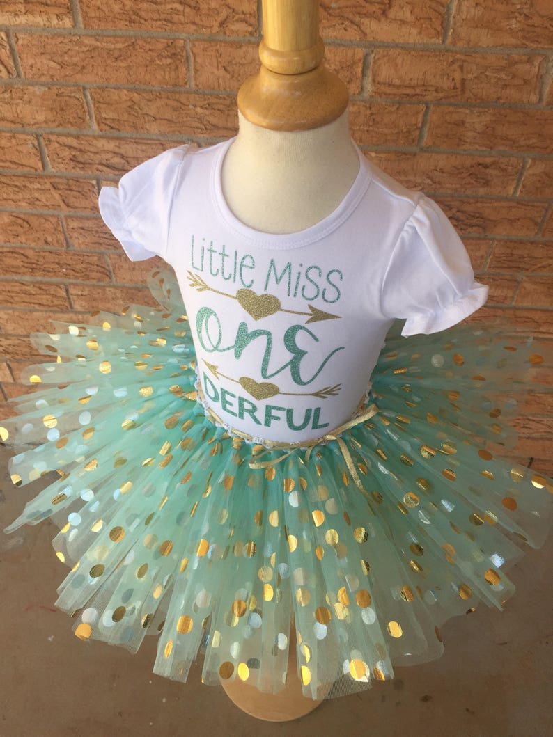 May include: A white t-shirt with the text "Little Miss One Derful" in gold glitter and a green arrow with a gold heart. The shirt is paired with a mint green tulle tutu with gold polka dots.