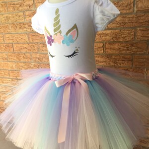 Peut inclure: Un t-shirt blanc avec un motif de visage de licorne orn&eacute; de paillettes dor&eacute;es, de fleurs roses et bleues et de cils en paillettes dor&eacute;es. Le t-shirt est associ&eacute; &agrave; un tutu en tulle arc-en-ciel avec un ruban de satin rose.