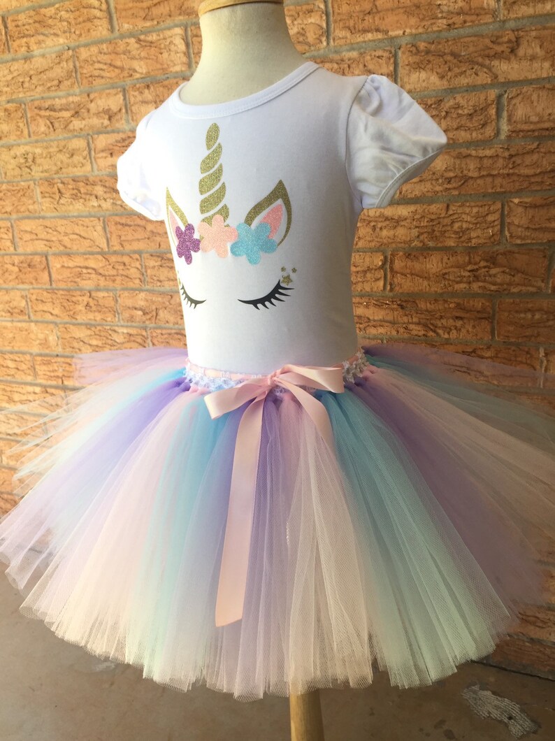 Peut inclure: Un t-shirt blanc avec un motif de visage de licorne orn&eacute; de paillettes dor&eacute;es, de fleurs roses et bleues et de cils en paillettes dor&eacute;es. Le t-shirt est associ&eacute; &agrave; un tutu en tulle arc-en-ciel avec un ruban de satin rose.