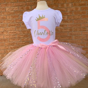 May include: A pink tulle tutu skirt with gold glitter dots and a white t-shirt with a pink number 5 and a gold crown, the text "Charlotte" is written below the number.