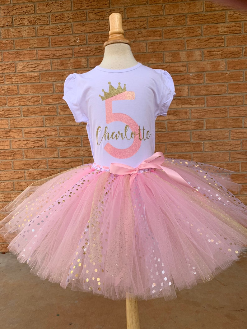 May include: A pink tulle tutu skirt with gold glitter dots and a white t-shirt with a pink number 5 and a gold crown, the text "Charlotte" is written below the number.