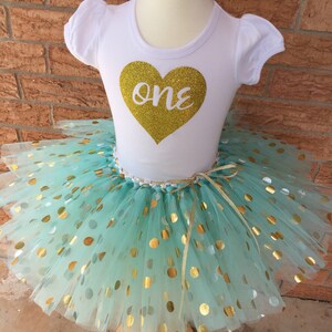 May include: A white t-shirt with a gold glitter heart that says "one" on it. The shirt is paired with a mint green tulle tutu with gold polka dots.