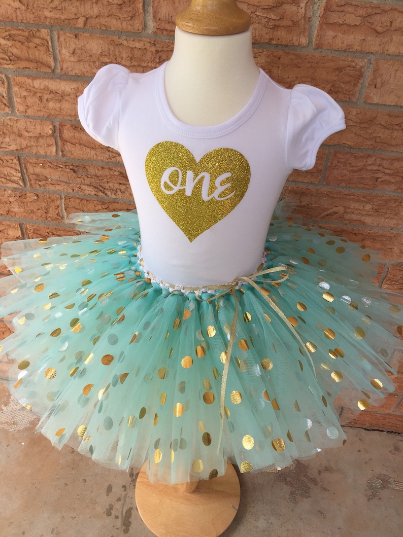 May include: A white t-shirt with a gold glitter heart that says "one" on it. The shirt is paired with a mint green tulle tutu with gold polka dots.