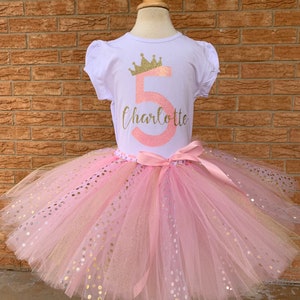 May include: A pink tulle tutu skirt with gold glitter polka dots. A white t-shirt with a gold crown and the number 5 in pink glitter with the name Charlotte underneath.