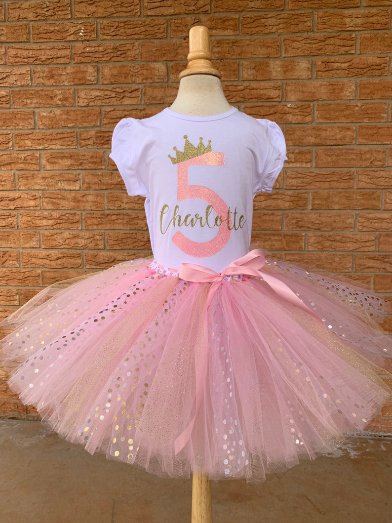 May include: A pink tulle tutu skirt with gold glitter polka dots. A white t-shirt with a gold crown and the number 5 in pink glitter with the name Charlotte underneath.