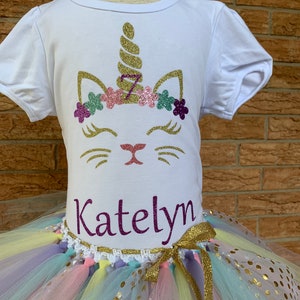 May include: White t-shirt with a gold glitter unicorn design and the name "Katelyn" printed on the bottom. The shirt is paired with a rainbow tulle tutu with gold glitter polka dots.