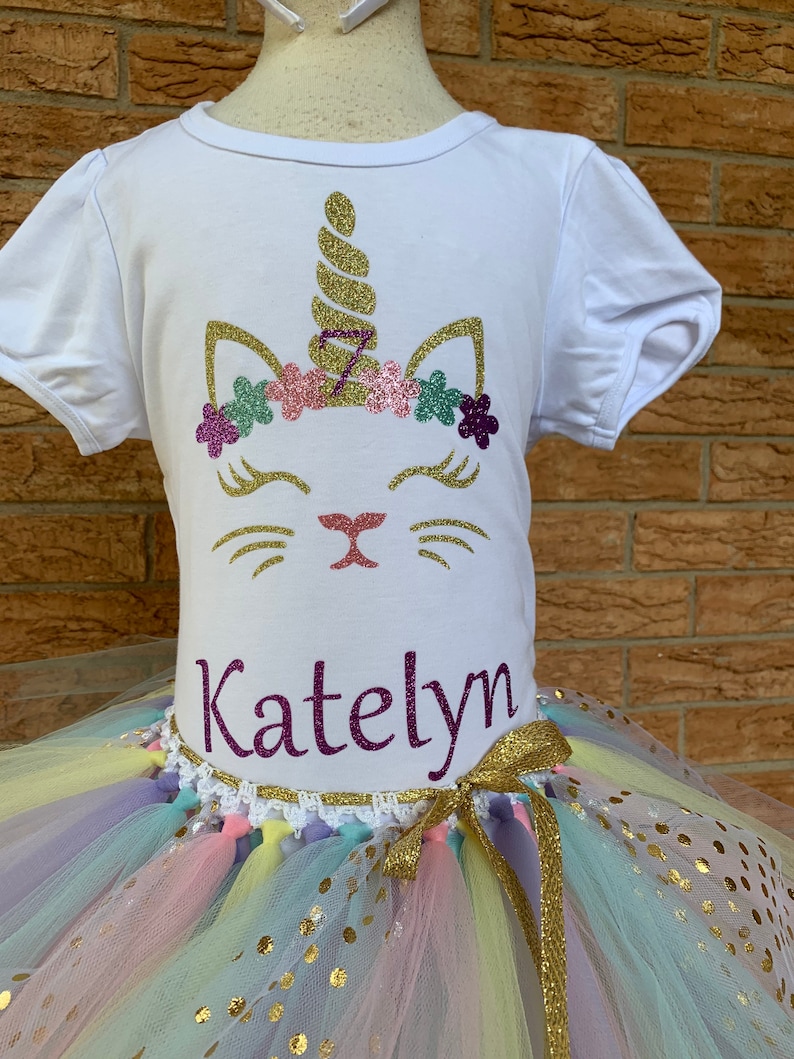 May include: White t-shirt with a gold glitter unicorn design and the name "Katelyn" printed on the bottom. The shirt is paired with a rainbow tulle tutu with gold glitter polka dots.
