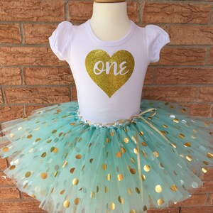 May include: A white t-shirt with a gold glitter heart that says "one" on it. The shirt is paired with a mint green tulle tutu with gold polka dots.
