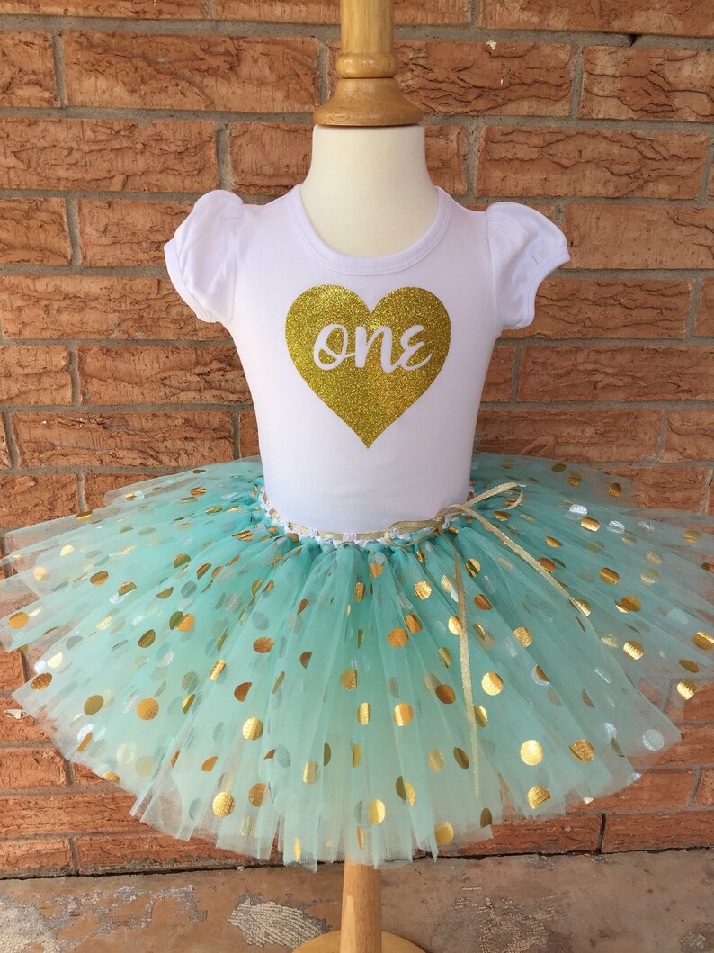 May include: A white t-shirt with a gold glitter heart that says "one" on it. The shirt is paired with a mint green tulle tutu with gold polka dots.