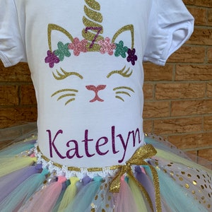 May include: A white t-shirt with a glittery unicorn cat design and the name "Katelyn" on the front. The shirt is paired with a rainbow tulle tutu with gold glitter polka dots.