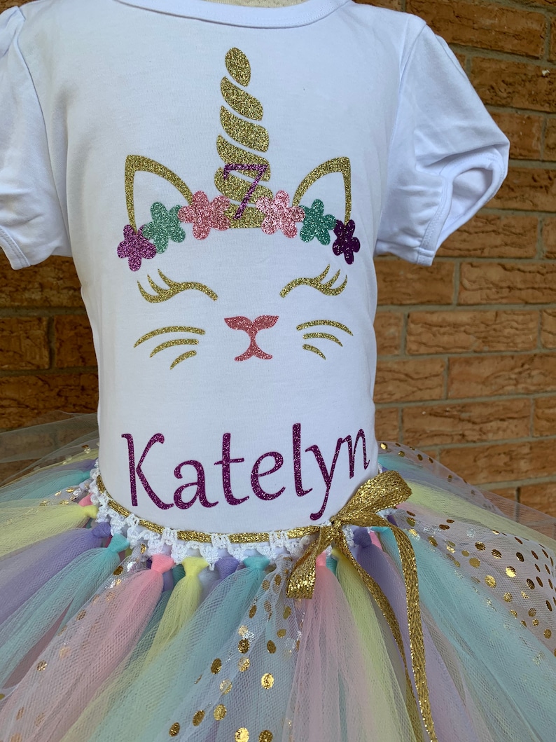 May include: A white t-shirt with a glittery unicorn cat design and the name "Katelyn" on the front. The shirt is paired with a rainbow tulle tutu with gold glitter polka dots.