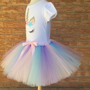 Peut inclure: Un t-shirt blanc avec un visage de licorne dor&eacute; et des fleurs sur le devant, associ&eacute; &agrave; une jupe tutu en tulle arc-en-ciel avec un ruban en satin rose.