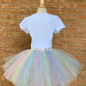 May include: A white t-shirt with a rainbow tulle tutu skirt with gold glitter dots. The skirt is layered and has a variety of colors, including pink, yellow, blue, and purple.