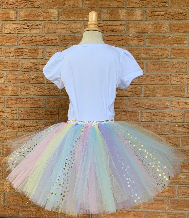 May include: A white t-shirt with a rainbow tulle tutu skirt with gold glitter dots. The skirt is layered and has a variety of colors, including pink, yellow, blue, and purple.