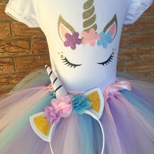Peut inclure: Une tenue de tutu de licorne blanche pour filles avec un t-shirt blanc repr&eacute;sentant un visage de licorne avec une corne dor&eacute;e et des fleurs roses, violettes et bleues. Le tutu est compos&eacute; de tulle superpos&eacute; dans des tons de violet, rose et bleu. La tenue comprend un bandeau blanc avec des oreilles paillet&eacute;es dor&eacute;es et une corne de licorne violette et blanche.