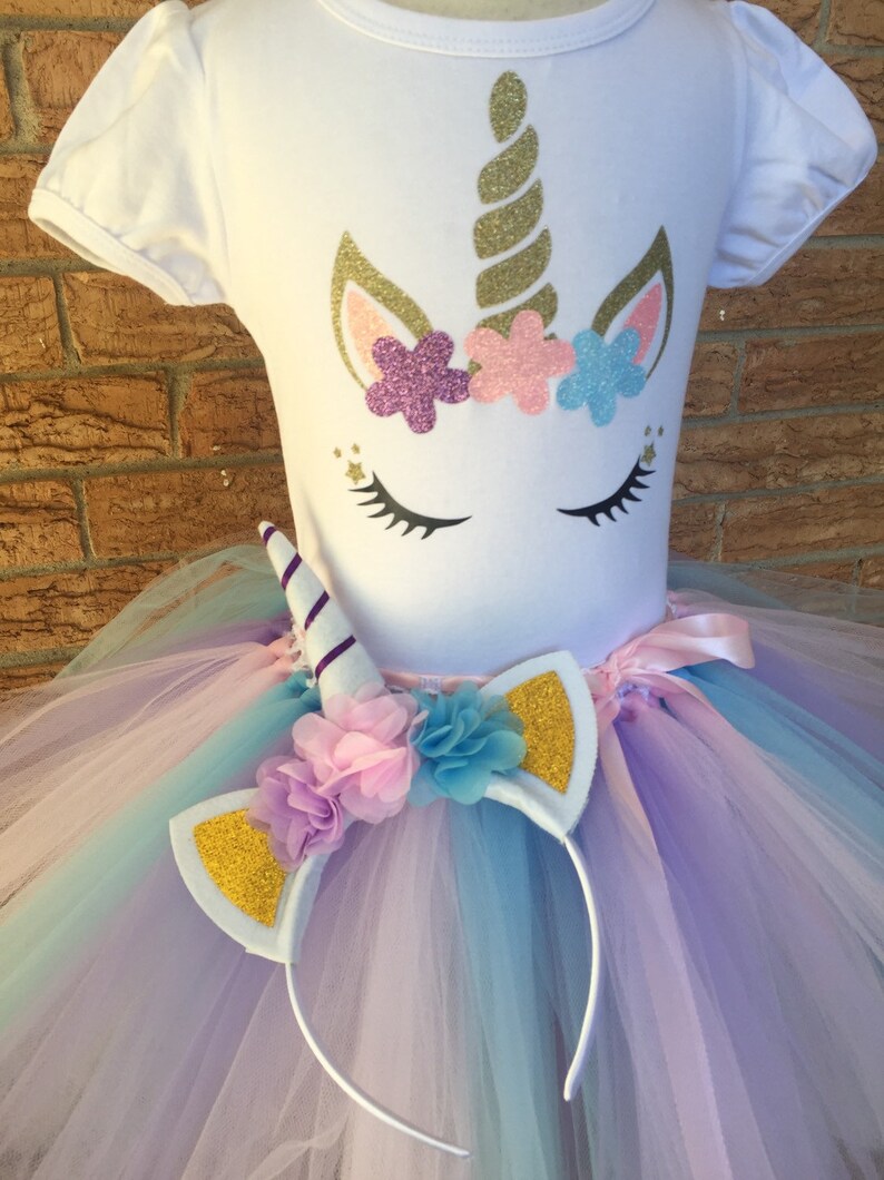 Peut inclure: Une tenue de tutu de licorne blanche pour filles avec un t-shirt blanc repr&eacute;sentant un visage de licorne avec une corne dor&eacute;e et des fleurs roses, violettes et bleues. Le tutu est compos&eacute; de tulle superpos&eacute; dans des tons de violet, rose et bleu. La tenue comprend un bandeau blanc avec des oreilles paillet&eacute;es dor&eacute;es et une corne de licorne violette et blanche.