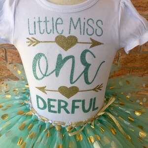 May include: A white onesie with the text "Little Miss One Derful" in teal and gold glitter. The onesie is paired with a mint green tulle skirt with gold polka dots.