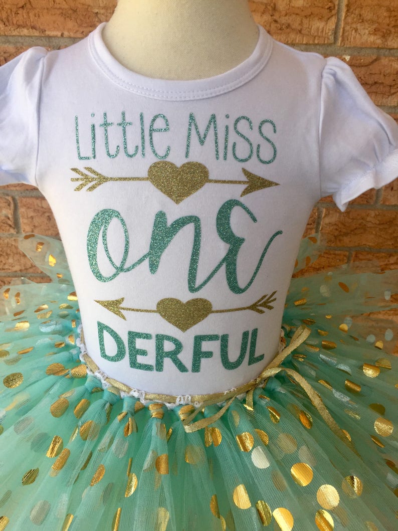 May include: A white onesie with the text "Little Miss One Derful" in teal and gold glitter. The onesie is paired with a mint green tulle skirt with gold polka dots.