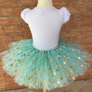 May include: A mint green tulle tutu skirt with gold polka dots. The skirt is layered and full, and the polka dots are scattered randomly.