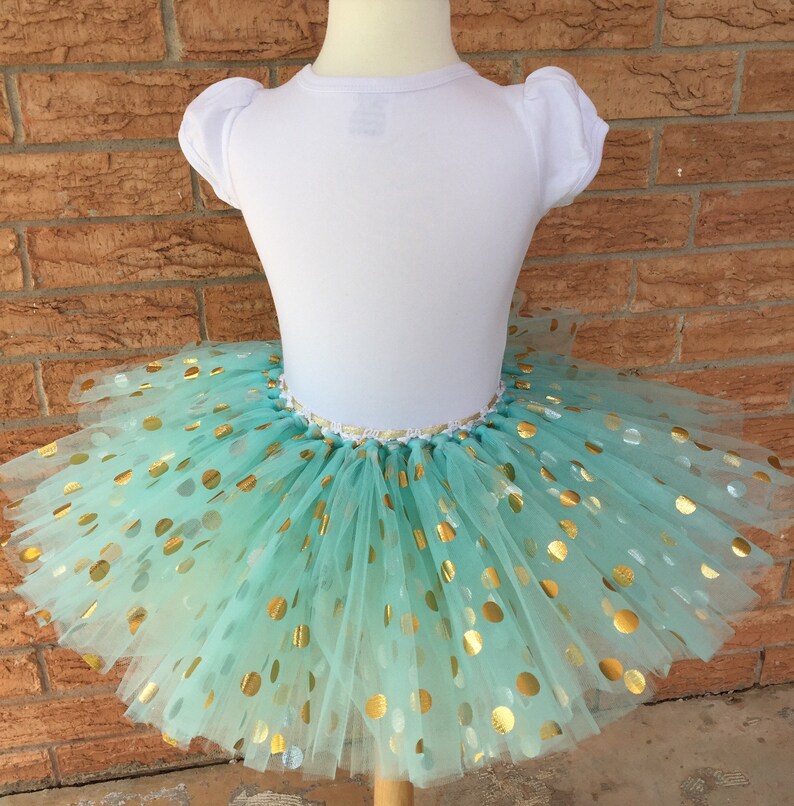 May include: A mint green tulle tutu skirt with gold polka dots. The skirt is layered and full, and the polka dots are scattered randomly.