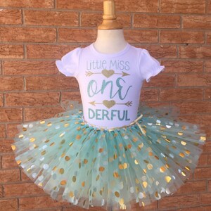 May include: A white t-shirt with the text "Little Miss One-derful" in gold and teal glitter. The shirt is paired with a teal tulle tutu with gold polka dots.