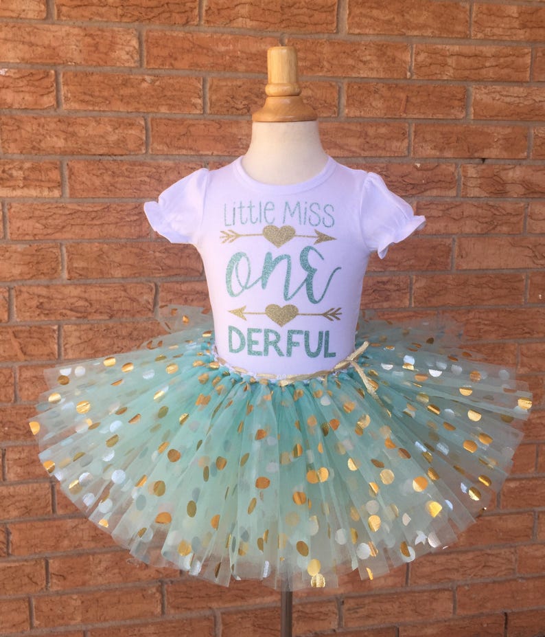May include: A white t-shirt with the text "Little Miss One-derful" in gold and teal glitter. The shirt is paired with a teal tulle tutu with gold polka dots.
