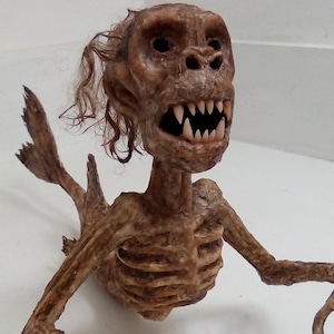 May include: A detailed sculpture of a creature with a skeletal torso, arms, and a fish-like tail. The creature has a brown, monkey-like face with sharp teeth and curly hair. The sculpture is set against a plain white background.