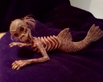 Könnte beinhalten: Detaillierte Skulptur einer skelettartigen Meerjungfrau mit Fischschwanz und knöchrigem Brustkorb. Die Figur hat ein humanoides Gesicht mit eingesunkenen Augen und spärlichem Haar. Die Meerjungfrau liegt auf einer dunkelvioletten Oberfläche.