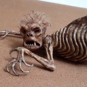 May include: A detailed sculpture of a creature with a skeletal torso and a primate-like head. The figure has sharp teeth, a fur-like mane, and outstretched arms with clawed hands. The color palette is primarily brown and tan.