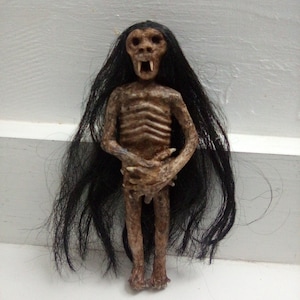 May include: A small, detailed figurine of a skeletal creature with long black hair. The figure has a gaunt face with visible teeth and is posed with its hands clasped. The color palette is primarily brown and black.